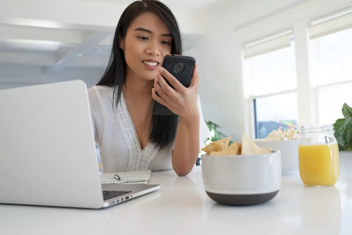 Young woman using laptop and smartphone while eating tortilla chips